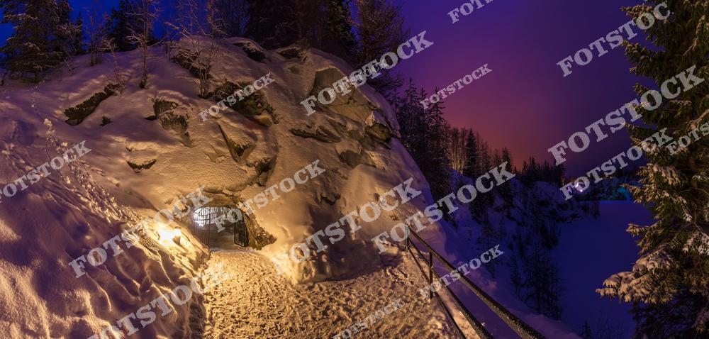 На фотографии запечатлён вход в одну из штолен горного парка «Рускеала» — известной достопримечательности Карелии. Зимний вечер окрашивает небо в фиолетово-розовые тона, а мягкое освещение придаёт маршруту загадочную атмосферу. Снег покрывает всё вокруг, подчёркивая суровую, но красивую природу Сортавальского района. Этот туристический маршрут популярен в зимний период, особенно во время рождественских и новогодних каникул.
